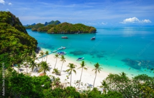 A tropical beach with white sand, palm trees, and clear turquoise water—perfect for Tailor-made Thailand Holidays. Boats are moored near the shore, with lush green islands in the background under a bright blue sky with wispy clouds.