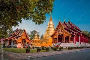 A golden pagoda and ornate wooden temples at Wat Phra Singh temple complex in Chiang Mai, Thailand, photographed at sunset with vibrant blue sky and lush greenery—perfect for unforgettable tailor-made Thailand holidays.