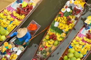 Three wooden boats filled with colourful fruits like bananas, pineapples, and pomelos float on a river. A vendor in a conical hat arranges produce—an authentic scene often featured on tailor-made Thailand holidays. The view is from above.