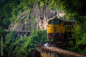 A yellow train travels along a curved railway track on a wooden bridge, surrounded by lush green trees and rocky cliffs—a scene reminiscent of the landscapes you’ll discover on tailor-made Thailand holidays in scenic, forested regions.