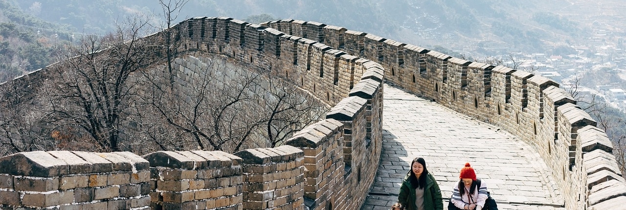 Three people, including a child, stroll along the stone pathway of the Great Wall of China on a sunny day, surrounded by mountains and bare trees—an unforgettable luxury holiday in Asia.