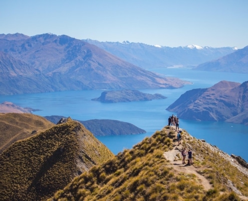 Walkers stroll along a narrow mountain ridge overlooking a blue lake and distant mountains under a clear sky, with grassy terrain in the foreground—an unforgettable scene on luxury New Zealand holidays.