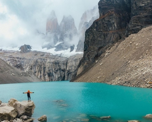 A person in an orange jacket stands on a rock at the edge of a bright turquoise lake, surrounded by steep rocky cliffs and misty, snow-capped mountains.