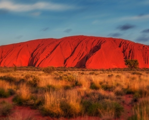 A large red sandstone rock formation, Uluru, rises from the flat, grassy desert under a blue sky with scattered clouds—an iconic sight often paired with Luxury New Zealand holidays in the awe-inspiring landscapes of the region.
