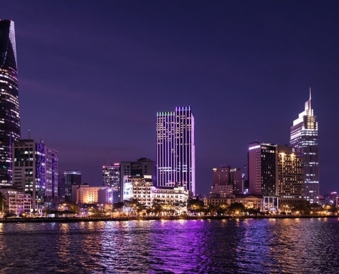 A vibrant city skyline at night with modern, brightly lit skyscrapers reflecting on a river under a clear purple-blue sky—perfect for capturing memories during your tailor-made Vietnam holidays.