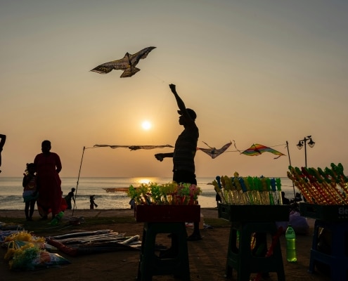A person enjoys flying a kite near the beach at sunset during Sri Lanka Holidays, with colourful toys displayed on tables in the foreground and silhouettes of others set against the vibrant sky and sea.