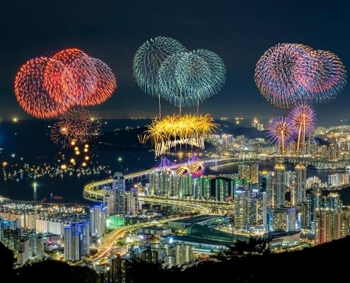 A night-time cityscape with tall buildings and a brightly lit bridge, illuminated by large, colourful fireworks exploding in the sky above the city during holidays in South Korea.