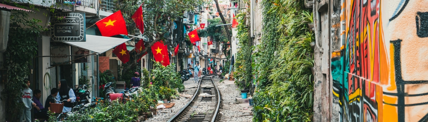 A narrow railway track runs through a lively urban street lined with buildings, greenery, and street art. Numerous Vietnamese flags hang above cafés and shops, offering a glimpse into daily life perfect for tailor-made Vietnam tours.