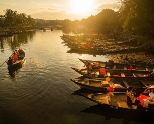 Boats with people wearing life jackets float on a calm river at sunset, golden light reflecting on the water—a picturesque moment often enjoyed during tailor-made Vietnam holidays. Trees and more boats line the shore beneath a partly cloudy sky.