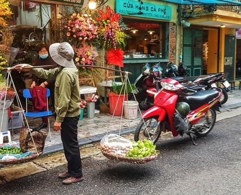 A street vendor balances baskets of produce on a bamboo pole outside a flower shop, with colourful bouquets, a red scooter, and parked motorbikes on a bustling city pavement—an authentic scene perfect for Tailor-made Vietnam Tours.