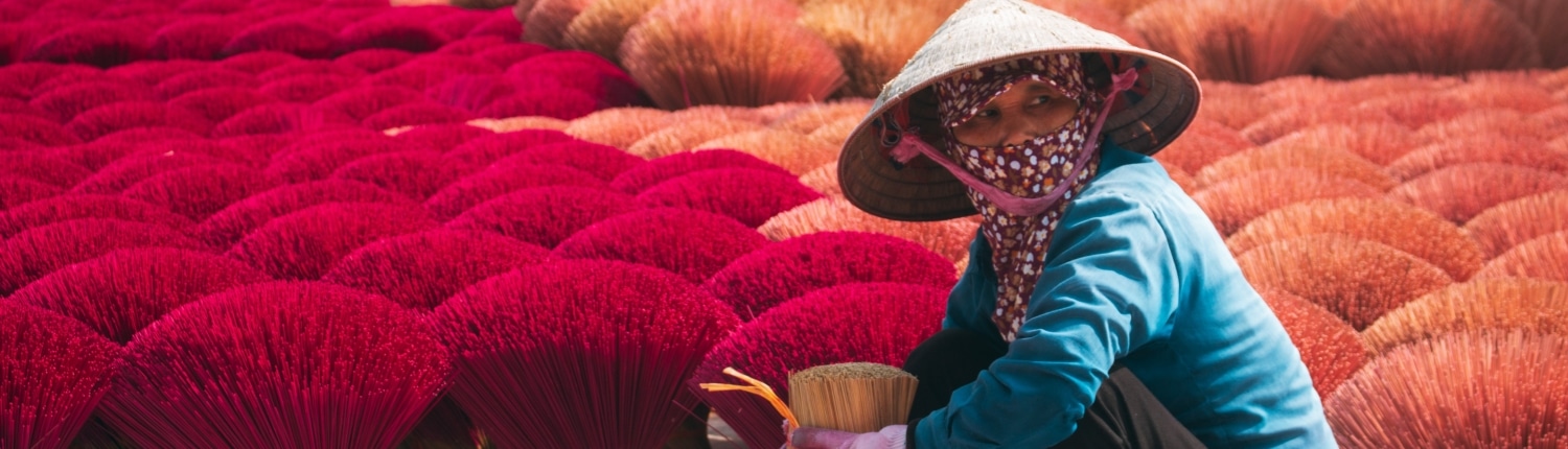 A person wearing a conical hat, mask, and gloves is squatting and sorting bundles of red incense sticks, surrounded by rows of drying incense—an authentic scene you can witness on tailor-made Vietnam holidays.