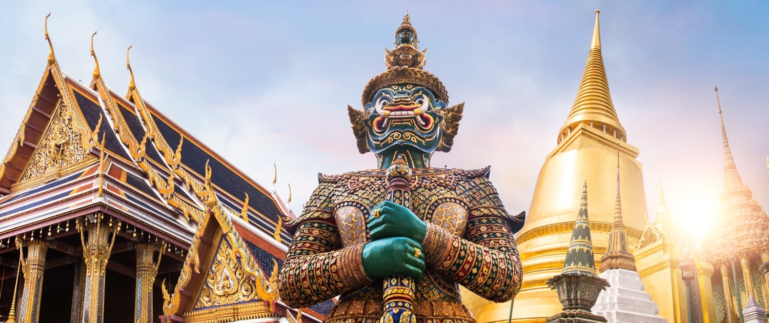 A colourful giant guardian statue stands before ornate temple buildings and a golden stupa at Wat Phra Kaew, Grand Palace, Bangkok—an iconic sight to discover on tailor-made Thailand holidays under a bright, sunlit sky.