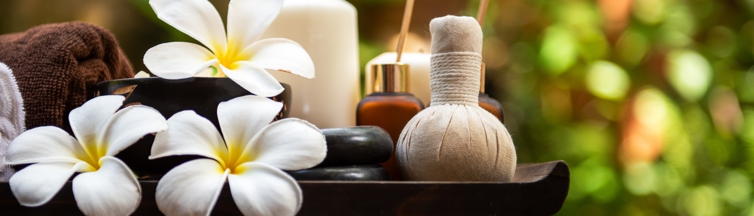 A spa setting with white frangipani flowers, stacked black stones, a herbal compress, essential oil bottles, incense sticks, a towel, and a lit white candle on a tray evokes the serenity of tailor-made Thailand holidays against a blurred green background.